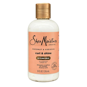 A bottle of SheaMoisture Curl & Shine Styling Milk in Coconut & Hibiscus, featuring an off-white bottle, a peach-colored label, and a metallic gold cap, set against a plain white background.