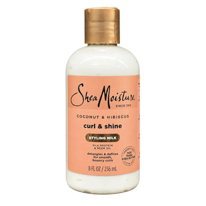 A bottle of SheaMoisture Curl & Shine Styling Milk in Coconut & Hibiscus, featuring an off-white bottle, a peach-colored label, and a metallic gold cap, set against a plain white background.